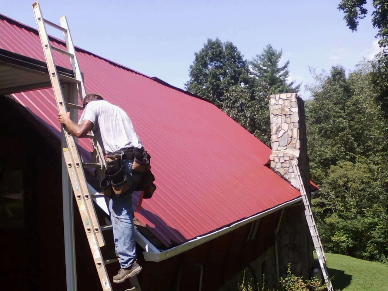 Roofer on a ladder beside a red metal roof and stone chimney — Soffit & Fascia Repair by Roofing Brothers in Brookpark